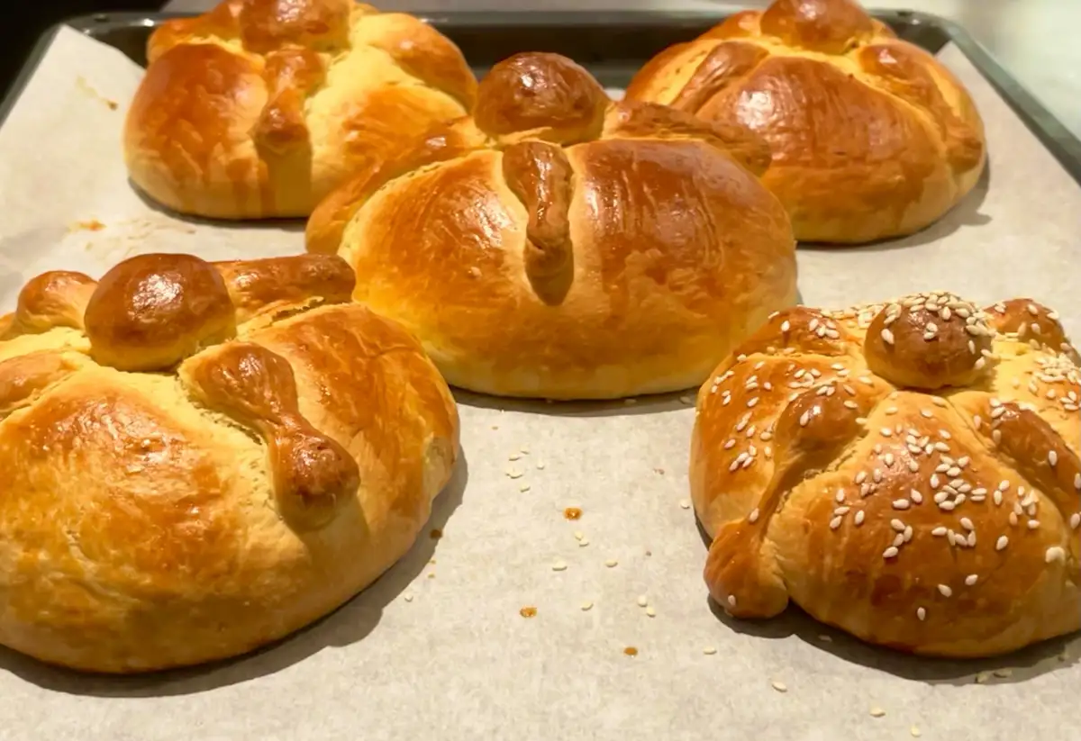 Five golden-brown, braided bread rolls sit on parchment paper on a baking tray. One roll in the foreground is topped with…