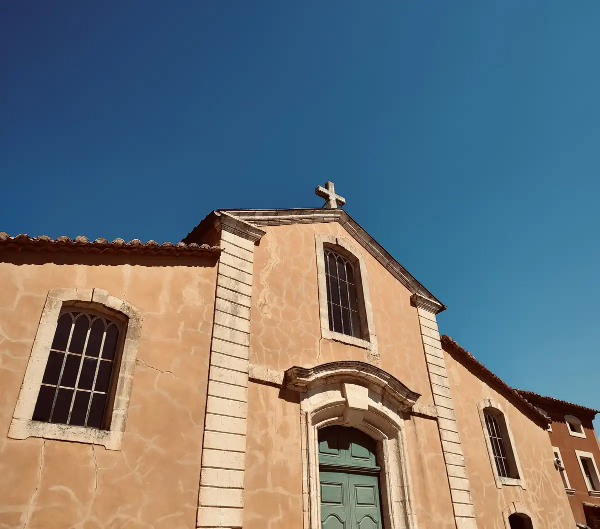 A low-angle photo of a peach-colored church with a green door and a cross on its roof against a clear blue sky. The overlaid…