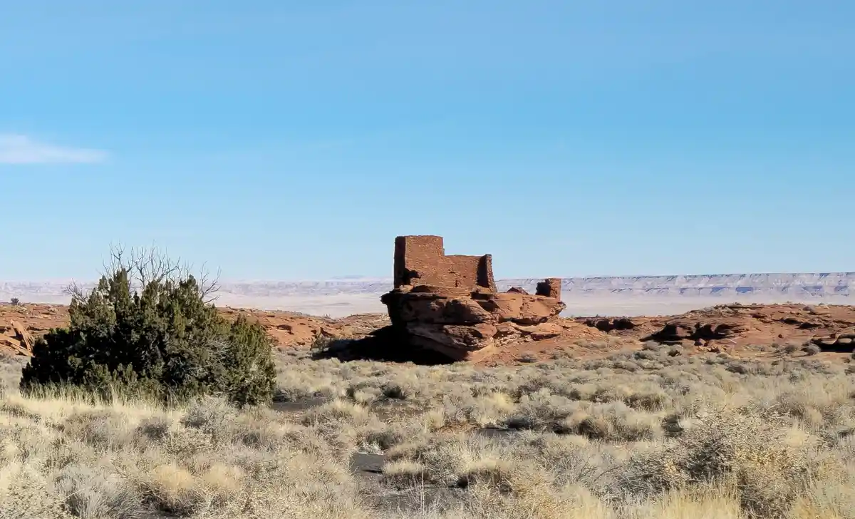 A wide desert landscape under a clear blue sky, featuring a prominent red rock formation with a small, weathered stone…
