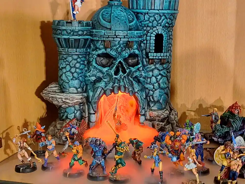 A collection of fantasy miniatures surrounds a large, skull-shaped castle with a glowing orange lava-like entrance. One…