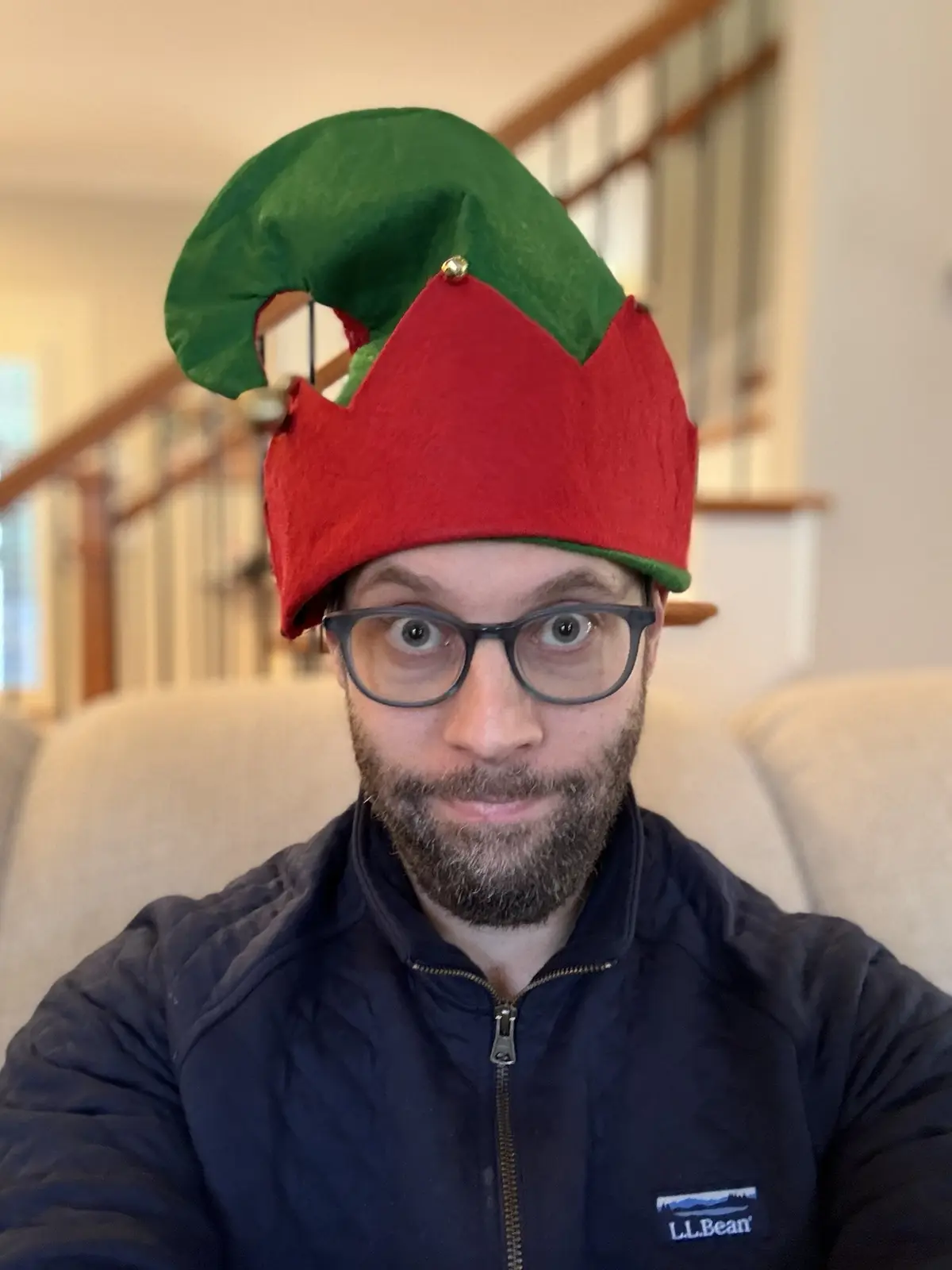 A man with glasses and a beard wears a red and green elf hat with a gold bell, looking directly at the camera with wide eyes…