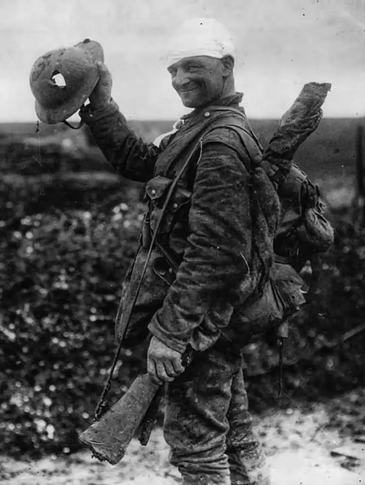 A black-and-white historical photo shows a smiling, muddy soldier with a bandaged head, holding up a damaged helmet with a…