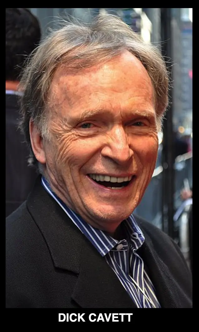 A close-up photo of an older man with gray hair, wearing a dark suit and striped shirt, smiling broadly with his mouth open…
