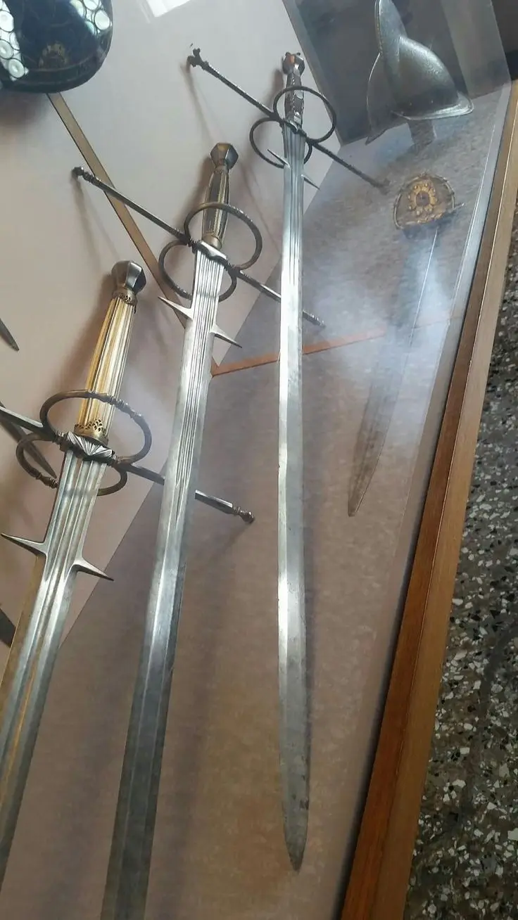 Three ornate historical swords are displayed vertically inside a glass case, with decorative hilts and crossguards. The…