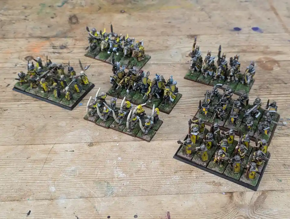 Several painted miniature wargaming armies, featuring knights, archers, and infantry in yellow and silver armor, are…