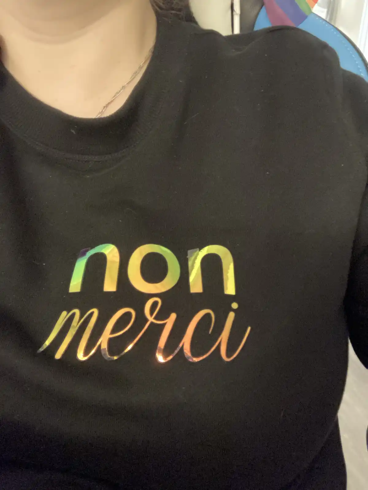 A close-up photo shows a person wearing a black sweatshirt with the words 'non merci' printed in shiny, iridescent…