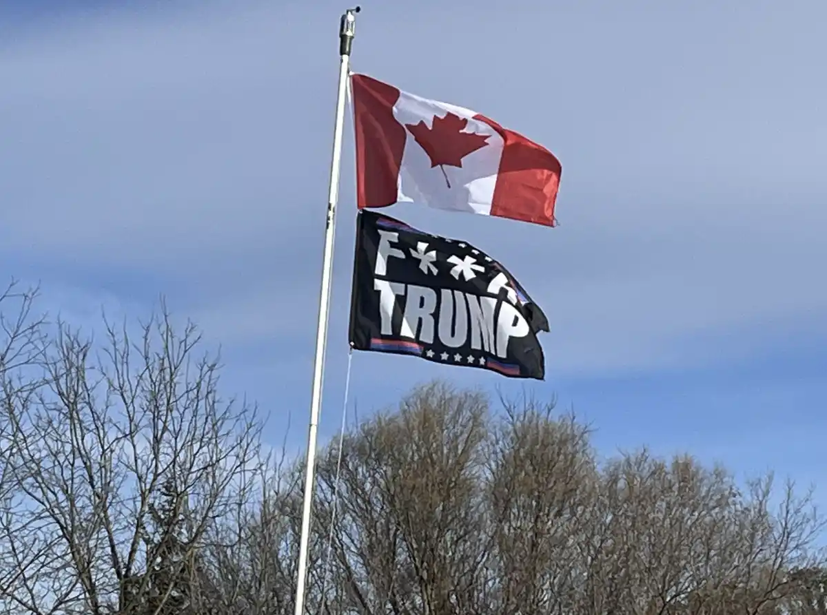 A Canadian flag flies above a black flag with white text reading 'F*** TRUMP' on a flagpole against a blue sky with bare…