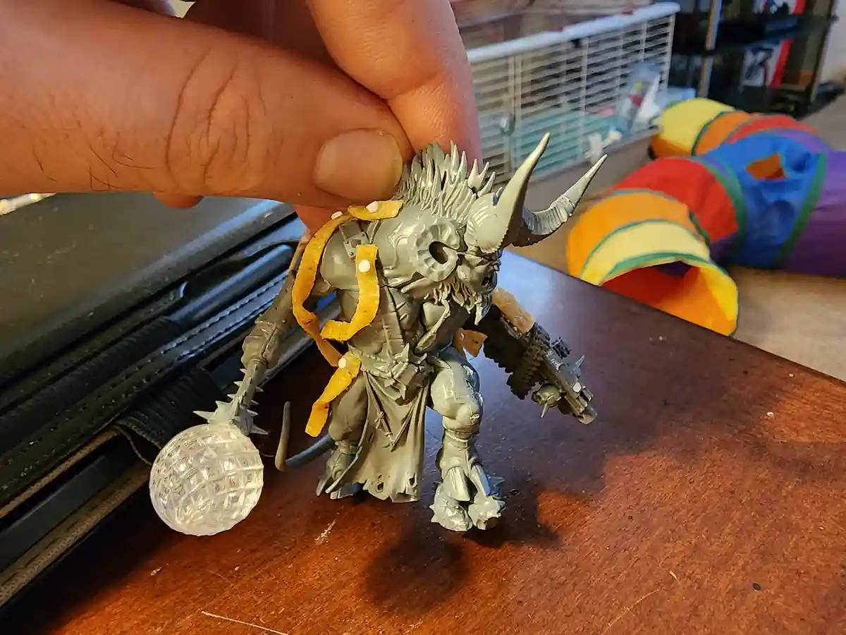 A hand holds a grey, unpainted fantasy miniature figure with horns and spikes, which has orange peel strips and white dots…
