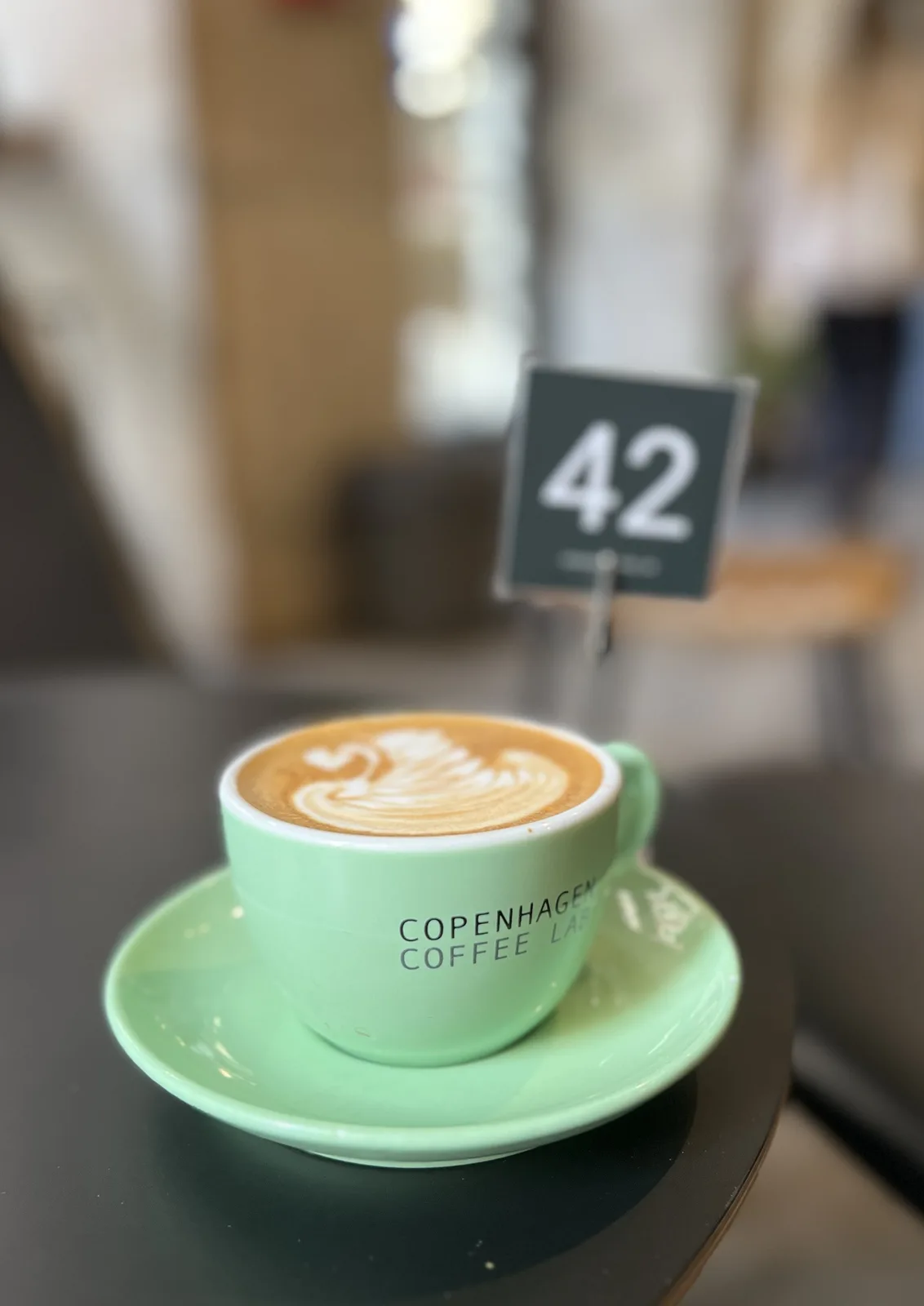 A mint green coffee cup with 'COPENHAGEN COFFEE LAB' printed on it sits on a matching saucer, featuring latte art on top…