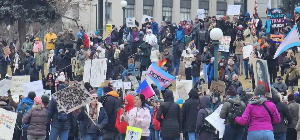 A large crowd of protesters in winter clothing holds various signs and flags, including a trans flag and signs with messages…