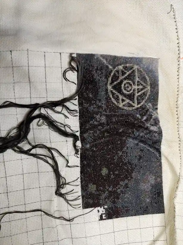 A close-up photo shows a partially completed cross-stitch project on white fabric with a grid pattern. Dark green embroidery…
