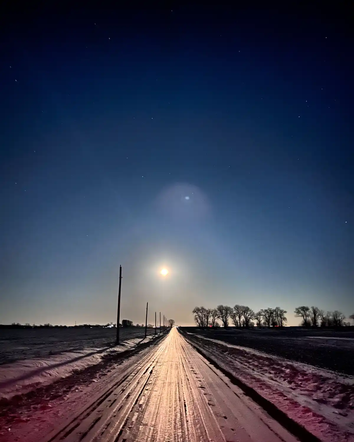 A snowy rural road stretches into the distance under a starry night sky, illuminated by a bright streetlamp and a visible…