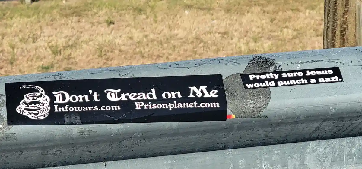 A close-up photo shows two stickers on a gray metal surface, likely a fence or railing, with dry grass in the background…
