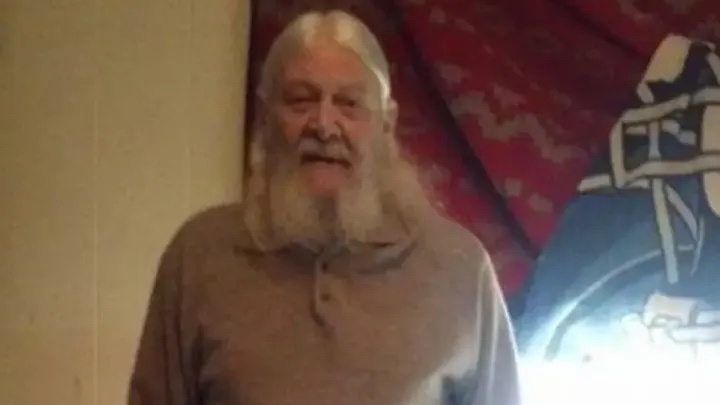 An older man with long white hair and a full beard, wearing a brown polo shirt, stands indoors in front of a red patterned…