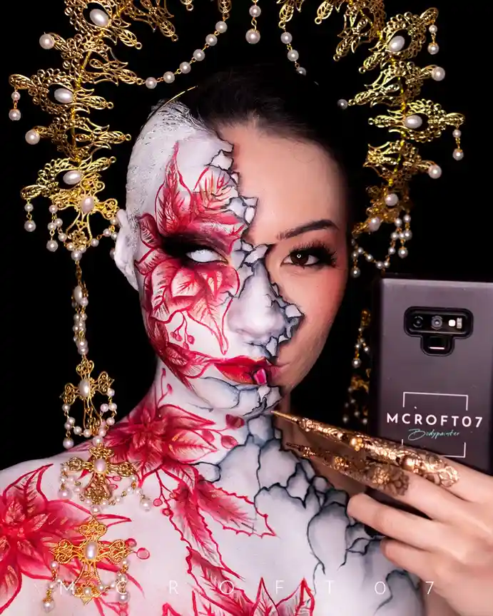 A person with elaborate body paint showing a split face: one side is a realistic human face with red floral designs, the…