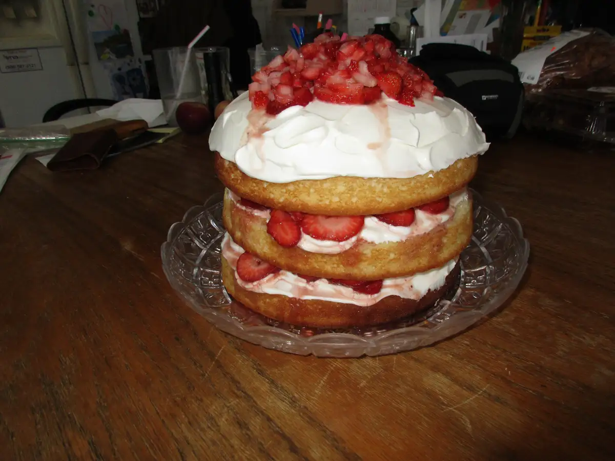 A three-layer strawberry shortcake sits on a clear glass plate atop a wooden table, with whipped cream and sliced…