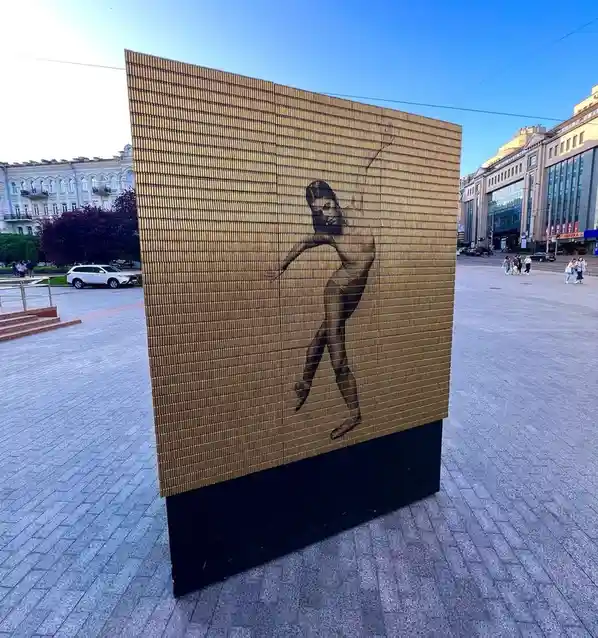 A large outdoor sculpture made of golden bullet casings arranged in a grid forms the silhouette of a nude woman dancing. The…