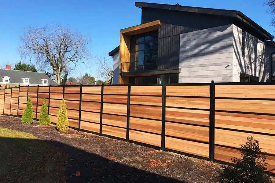 A modern wooden fence with black metal posts runs along the front of a contemporary house under a clear blue sky. The fence…