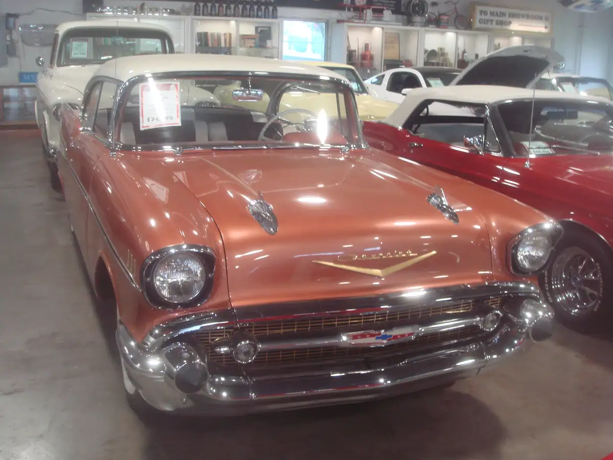 A shiny, rust-orange 1957 Chevrolet Bel Air is prominently displayed in a classic car showroom, with other vintage cars…