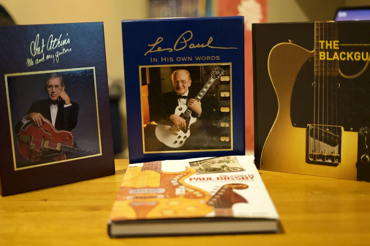 Four guitar-related books are arranged on a wooden surface. From left to right: 'Chet Atkins: Me and My Guitars' with a…