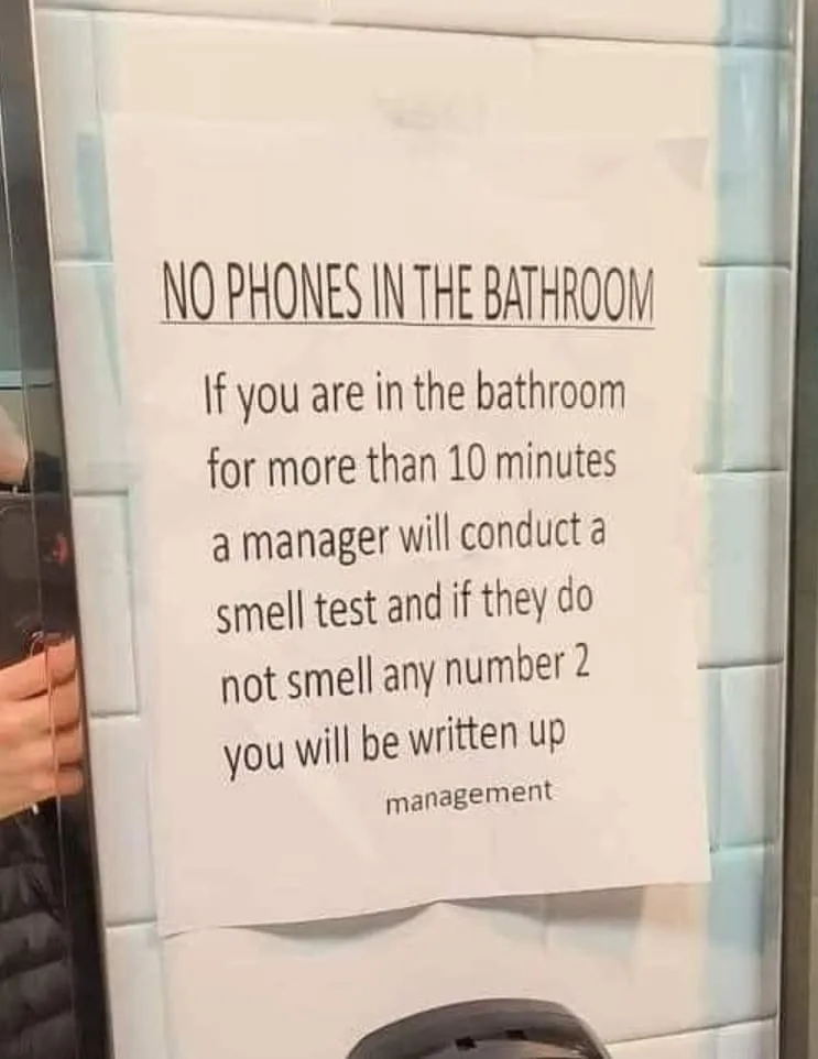 A white paper sign taped to a glass door in a tiled bathroom, with black text stating a rule about no phones and a humorous…
