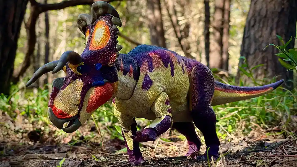 A colorful, toy triceratops dinosaur figurine with purple, yellow, and orange patterns stands in a forest setting. The…