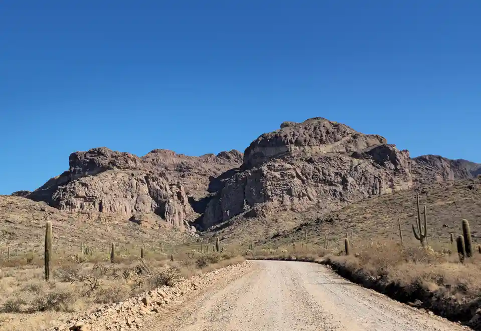 A wide-angle desert landscape photo shows a gravel road winding toward rugged, rocky mountains under a clear blue sky…