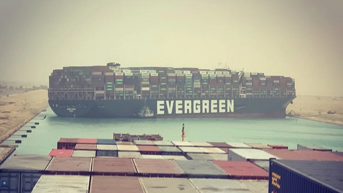 A massive container ship named 'EVERGREEN' is stuck sideways in the Suez Canal, blocking the waterway. In the foreground…