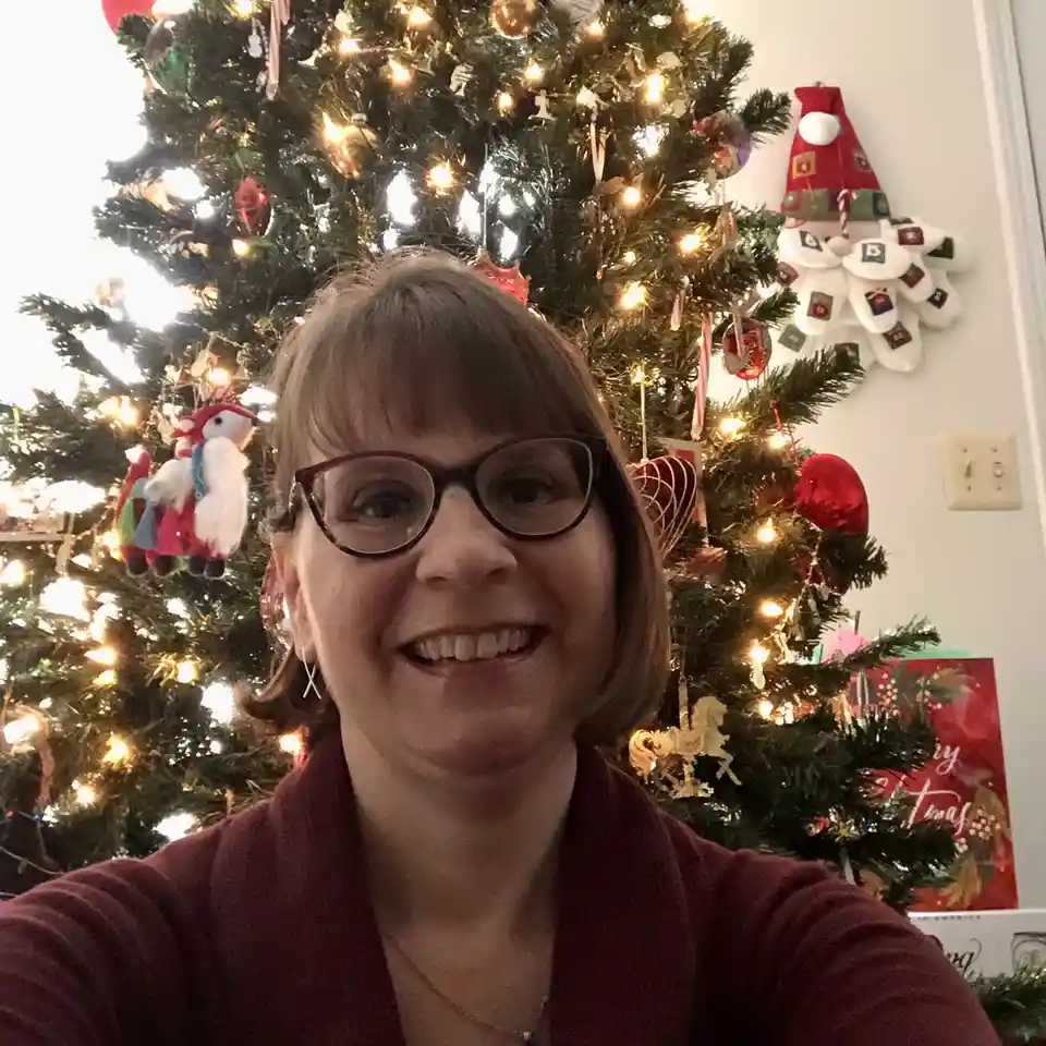 A woman with glasses and a red sweater smiles at the camera while standing in front of a brightly lit, decorated christmas…