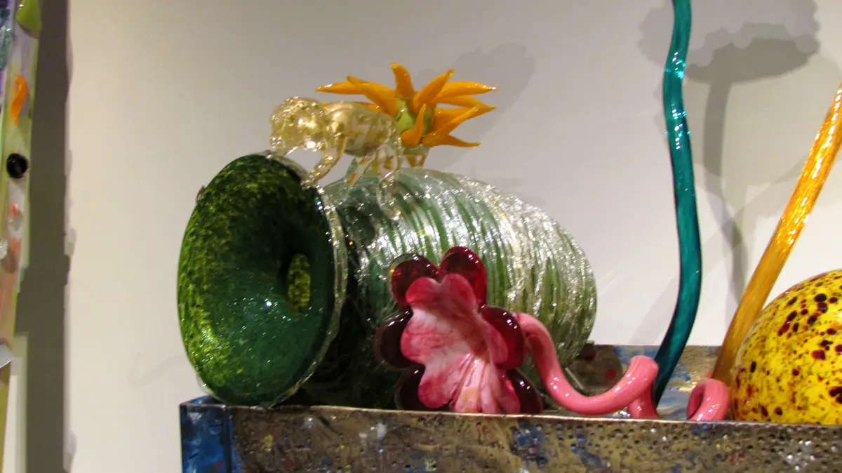A colorful glass sculpture features a green, ribbed, vase-like form with a translucent golden animal figurine perched atop…