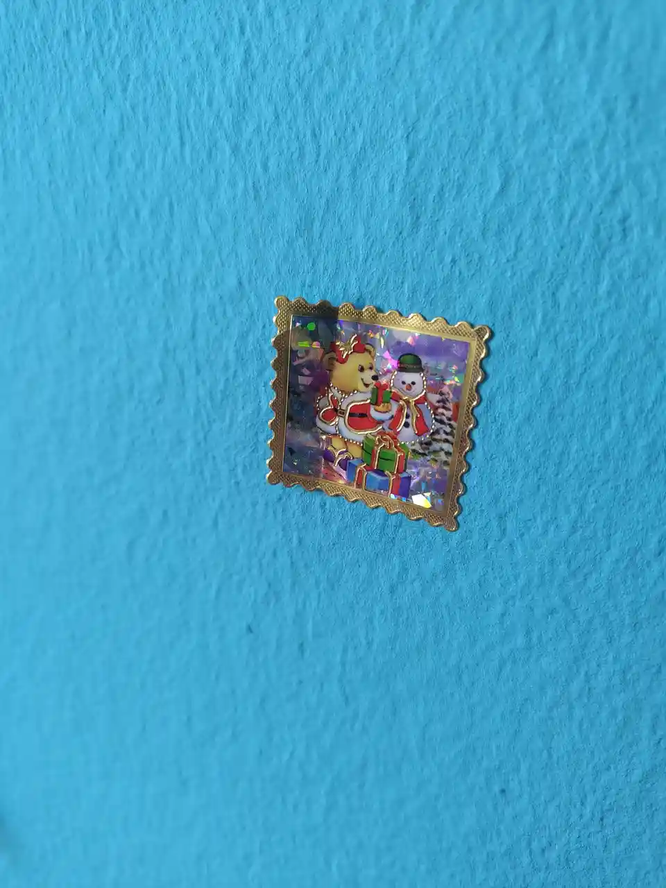 A small, square, gold-framed Christmas-themed pin with a holographic background sits on a textured blue surface. The pin…