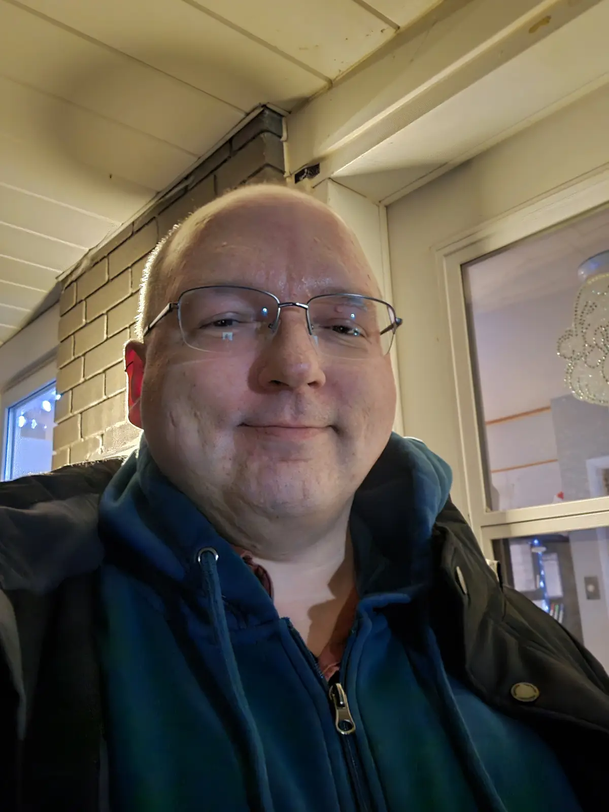 A close-up selfie of a bald man with glasses, wearing a blue hoodie under a dark jacket, standing in a doorway with brick…