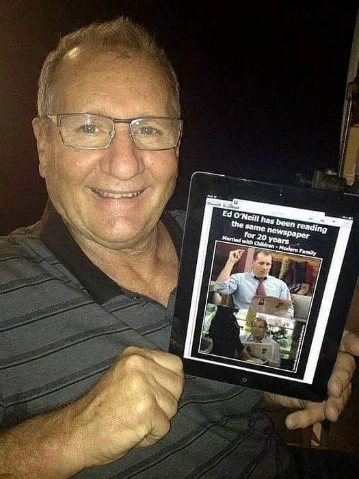 A smiling older man with glasses holds up a tablet displaying a news article about actor Ed O'Neill. The article's headline…