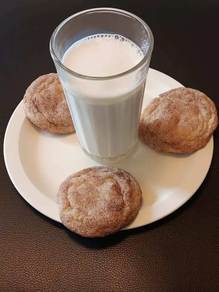 A white plate holds three cinnamon sugar cookies and a glass of milk on a dark brown textured surface. The overlaid text…