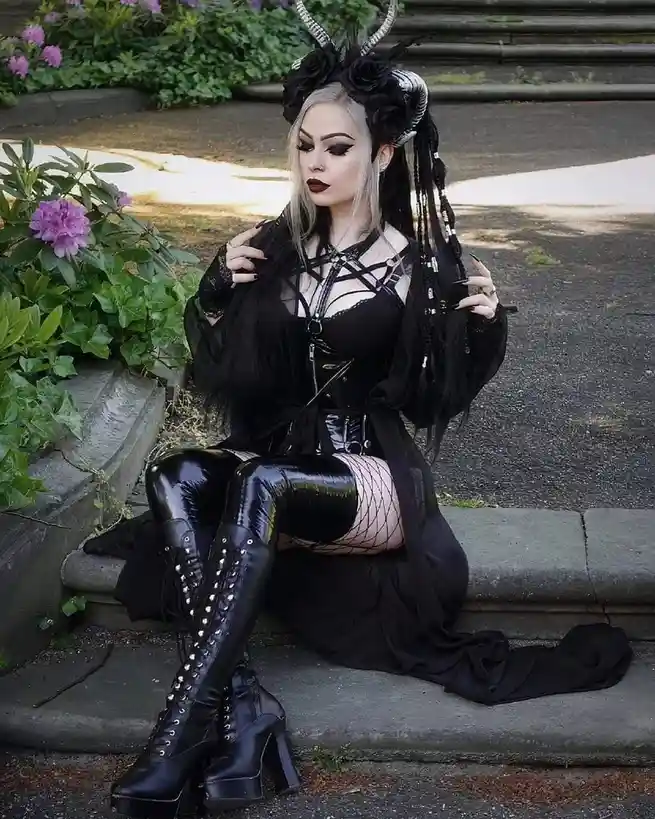 A woman with blonde hair and dark makeup sits on stone steps outdoors, wearing a black gothic outfit with horns, fishnet…