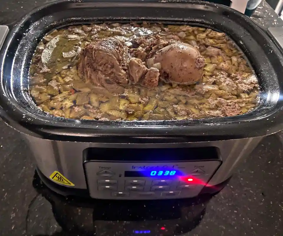 A close-up photo of a slow cooker filled with meat, onions, and broth, with the digital display showing '03:38' and a red…