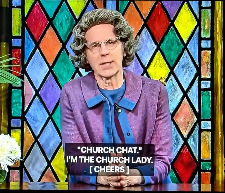 A person with gray curly hair and glasses, wearing a purple jacket over a blue shirt, sits in front of a colorful…