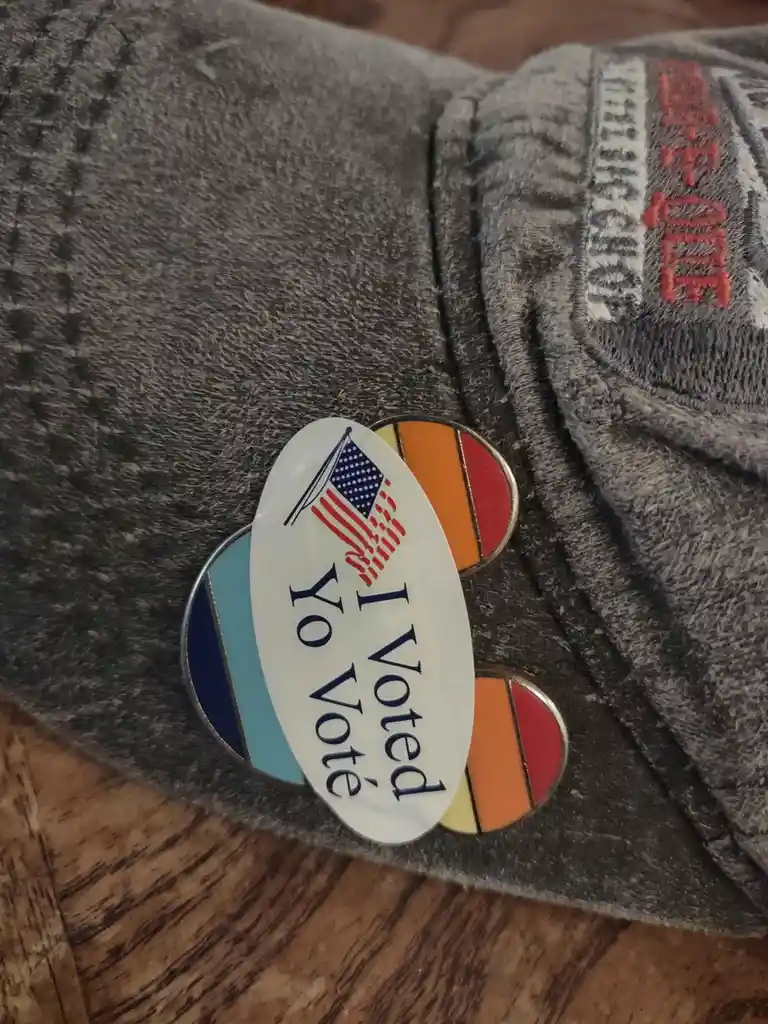 Three pins are pinned to a gray sweatshirt sleeve: a white oval pin with an American flag and the text 'I Voted Yo Vote', a…