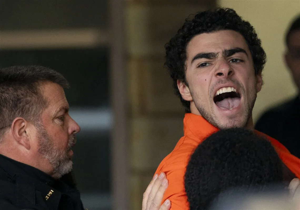 A man in an orange jumpsuit is being restrained by a uniformed officer while screaming with an intense, angry expression…