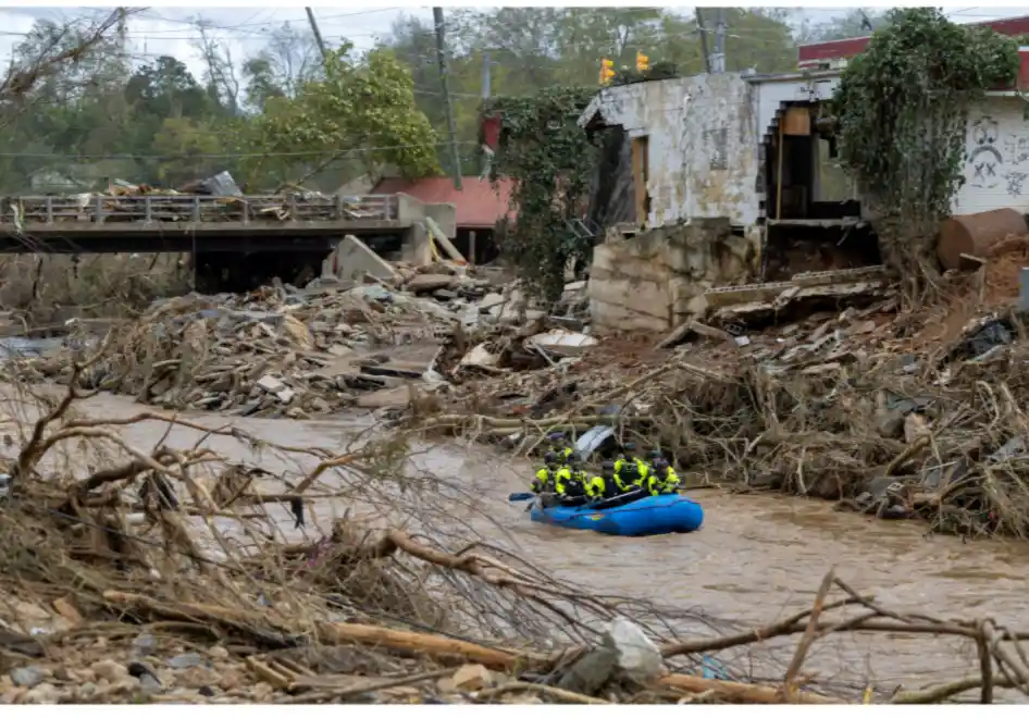 A photo shows several emergency responders in a blue inflatable raft navigating muddy, debris-filled floodwaters…