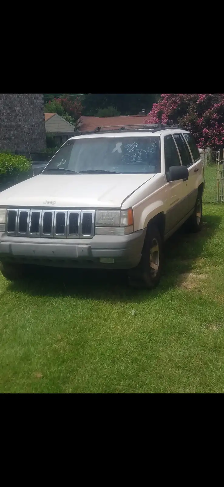 A white 1990s Jeep Grand Cherokee is parked on a grassy lawn with a house and flowering bushes in the background…