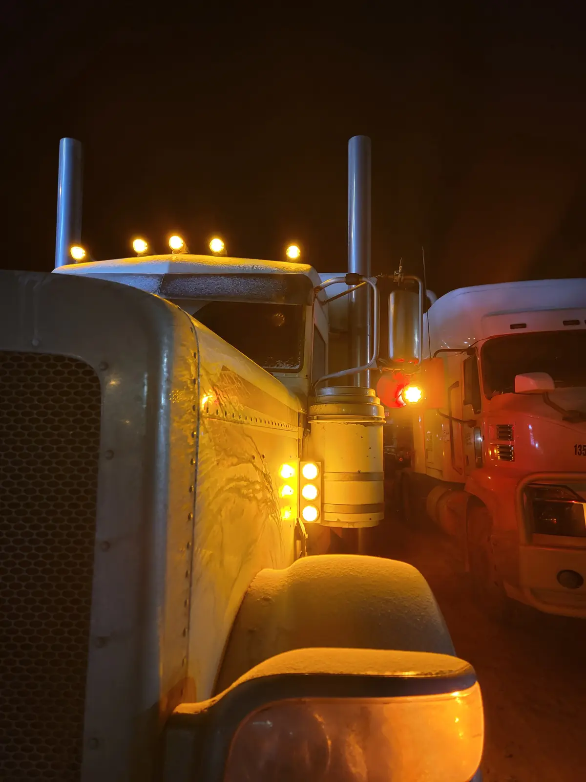 A close-up night photo shows two large semi-trucks parked side-by-side, their surfaces covered in snow and ice. The truck in…