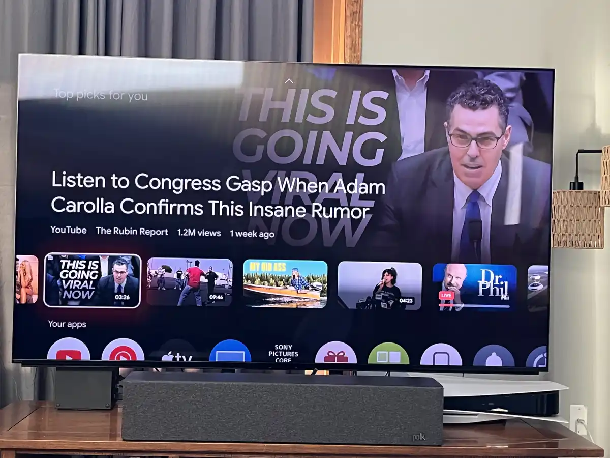 A television screen displays a YouTube video suggestion titled 'Listen to Congress Gasp When Adam Carolla Confirms This…