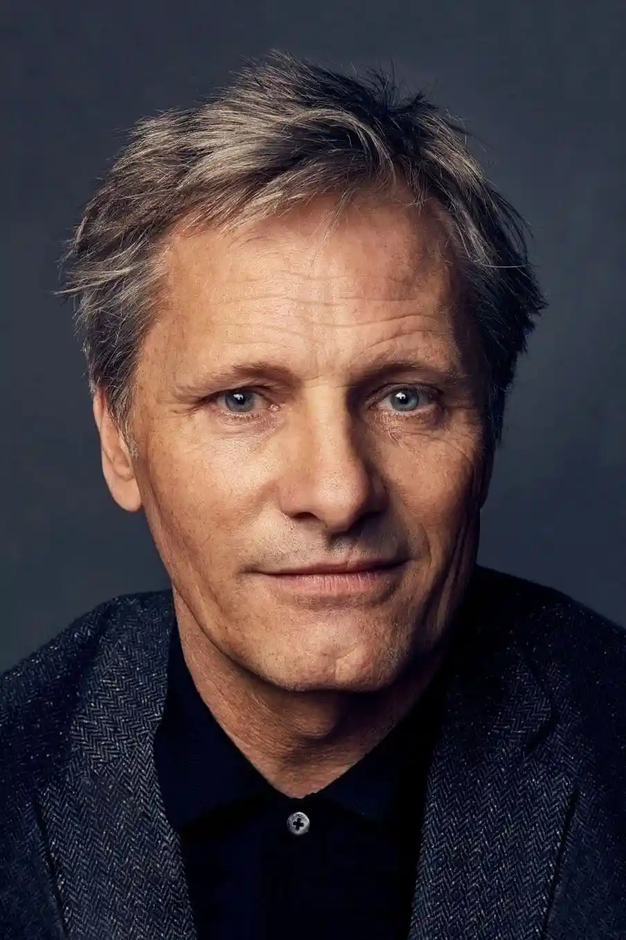 A close-up portrait of a middle-aged man with graying hair, blue eyes, and a subtle, knowing smirk. He wears a dark…