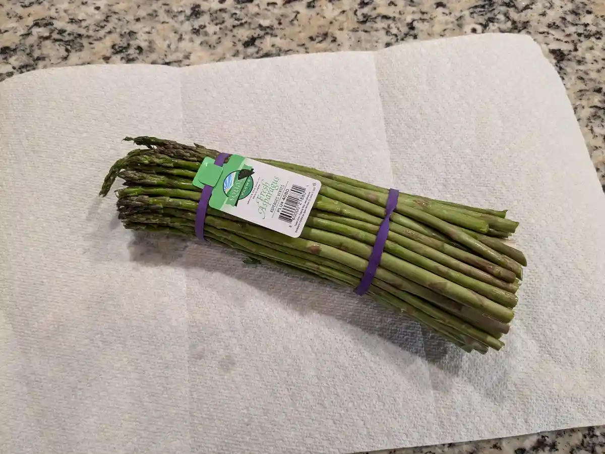 A bundle of fresh green asparagus spears, tied with a purple band and bearing a 'Squash Farms' label, rests diagonally on a…