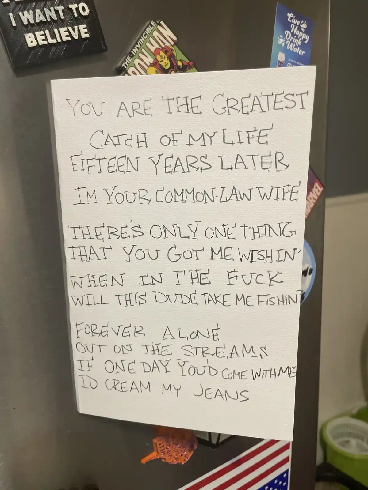 A handwritten note on white paper is stuck to a stainless steel refrigerator, surrounded by various magnets including 'I…
