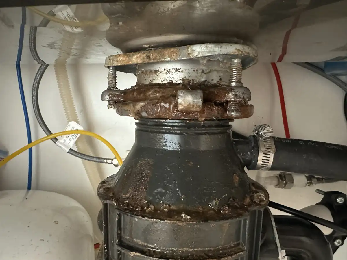 A close-up photo shows a severely corroded and grimy garbage disposal unit under a sink, with brown gunk and rust visibly…