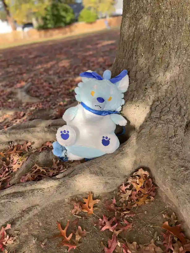 A light blue and white plush toy resembling a wolf or fox with blue paw prints and a blue bow sits nestled in the roots of a…