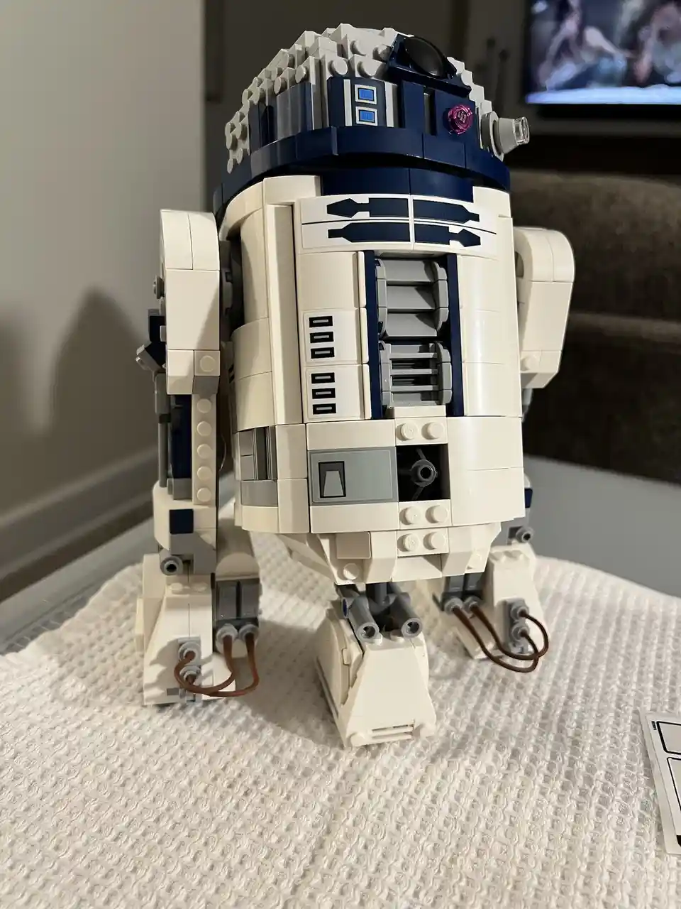 A detailed LEGO model of the Star Wars droid R2-D2 stands upright on a textured white surface, its iconic white and blue…