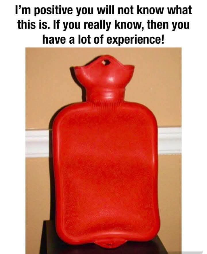 A red, textured hot water bottle with a rounded neck and a small hole at the top sits on a dark surface against a beige wall…